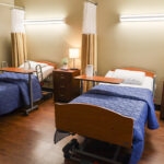 A room at the Annandale Healthcare Center with two twin beds featuring deep blue coverlets, a nightstand with a lamp and two drawers, and a dividing curtain.