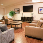 A common area at Annandale Healthcare Center featuring four armchairs and a coffee table with a tv mounted on the wall next to a fern and in between two pieces of art.