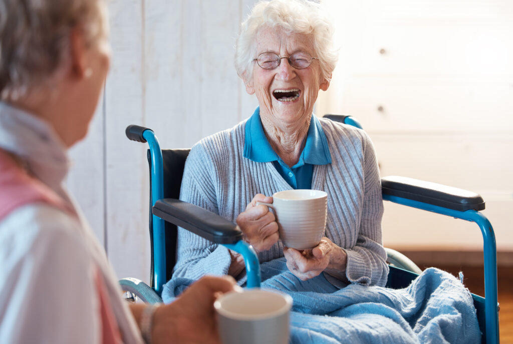 senior woman seated in a wheelchair and laughing with a friend while holding a mug.